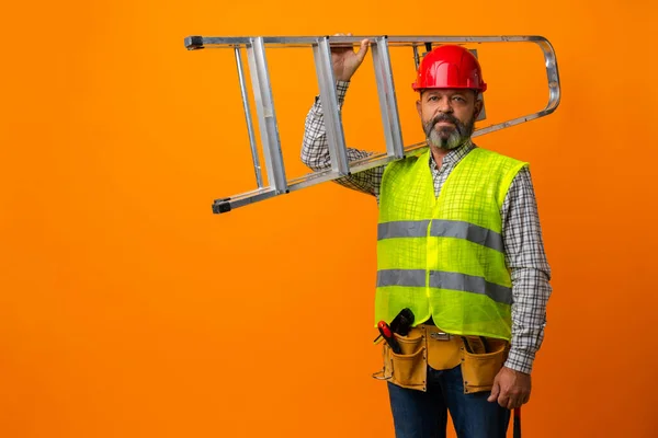 Handyman in uniform standing with ladder against orange background — Stock Photo, Image