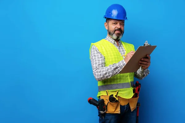 Construtor masculino ou faz-tudo em uniforme segurando prancheta contra fundo azul — Fotografia de Stock