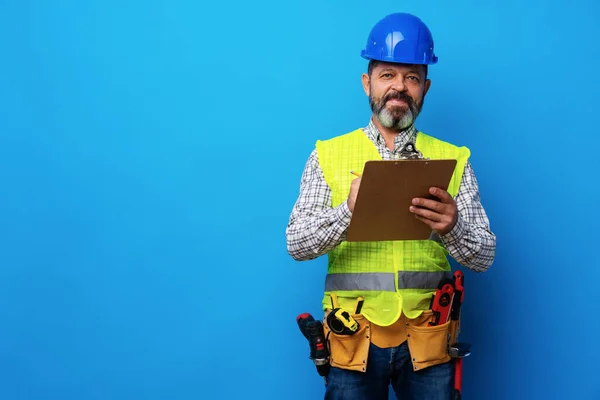 Construtor masculino ou faz-tudo em uniforme segurando prancheta contra fundo azul — Fotografia de Stock