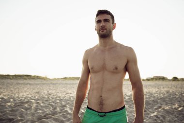 Gün doğumunda boş sahilde duran formasız genç adam.