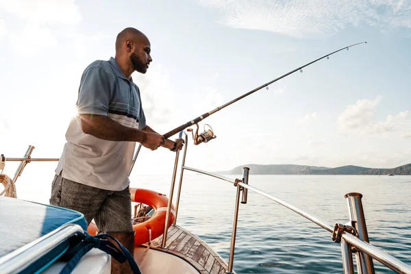Genç Afro-Amerikalı adam elinde oltayla bir yelkenli ile gün batımında açık denizde balık tutuyor. — Stok fotoğraf