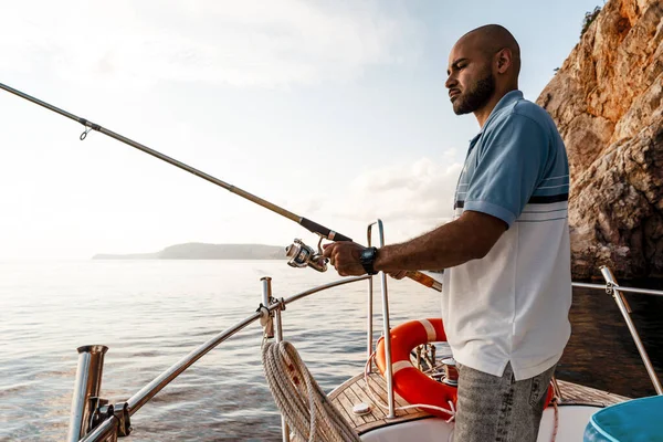 Junger afrikanisch-amerikanischer Mann steht mit Angelrute auf einem Segelboot beim Angeln im offenen Meer bei Sonnenuntergang — Stockfoto