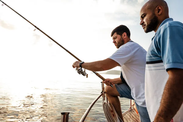 Deux jeunes hommes debout sur un voilier avec canne à pêche regardant haul — Photo