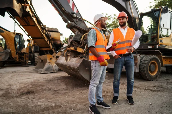 Två män ingenjörer diskuterar sitt arbete stående mot byggmaskiner — Stockfoto
