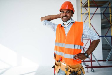 Sağlık maskesi takan, iş kıyafetleri ve şapka giyen melez ırk müteahhidinin portresi.