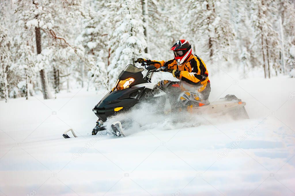A man in the outfit of a racer in a yellow-black overalls and a red-black helmet, driving a snowmobile at high speed riding through deep snow against the background of a snowy forest.