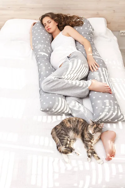 Beautiful sleeping pregnant woman with a big tummy with long curly hair and closed eyes, hugging a long body pillow with her arm and legs and lying on the bed with domestic cat indoor.