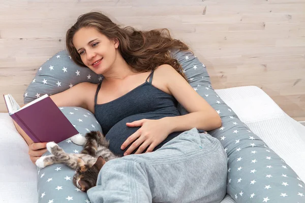 Beautiful long haired pregnant woman lying on bed on body pillow indoor with domestic cat, reading book and touching her big abdomen.
