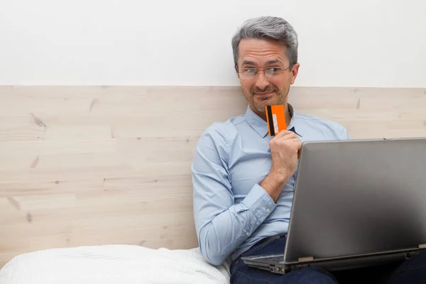 Middle-aged glad and interested man with grey hair wearing spectacles and sitting on the bed and working on a laptop and holding a banking credit card in your hand.