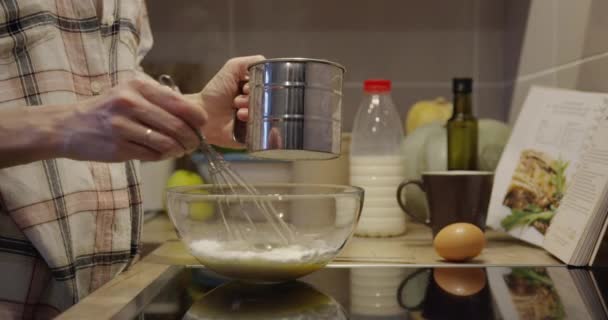 人間の手はレシピに従って食べ物を準備し 泡立て器で生地を混合 屋内食品とキッチンテーブルの背景にボウルに小麦粉を注ぐ — ストック動画