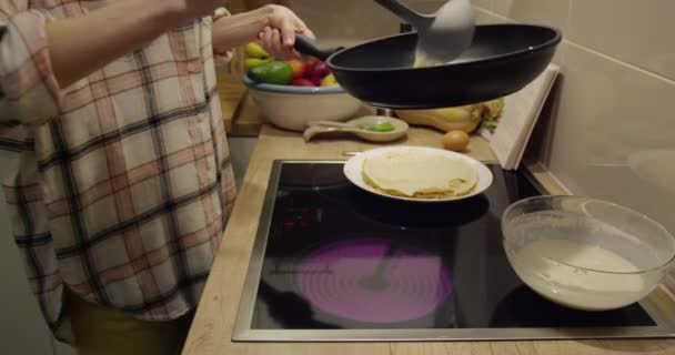 Human hands prepare pancakes, pour dough from a ladle into a hot frying pan, forming a pancake and fry it on the backdrop of a kitchen interior and a table with food indoors. — Stock Video