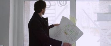 Mimarın elinde bina planı var. Arka plan panoramik penceresinde inşaat halindeki bir kır evinin planlarını izliyor ve kontrol ediyor..