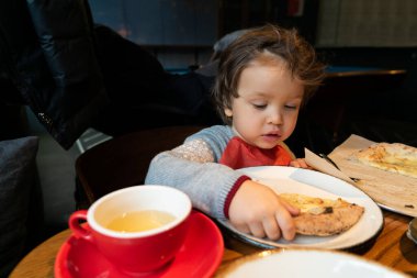 Küçük çocuk içeride masada oturuyor ve bir dilim pizzayı eliyle yiyor.