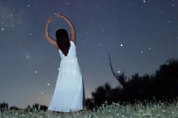 Woman under starry night in white long dress ballet pose raising arms Woman under night sky, Blurred night sky. Ballet pose under starry night.