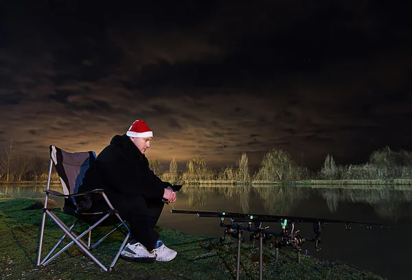 Pescador com chapéu de Papai Noel, Vinha na mão, sentado na cadeira olhando em varas, paciência Bela clodudscape à noite — Fotografia de Stock