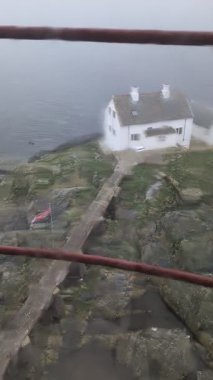 Yüksek açılı manzara, denizin yanında kalın gri sisle kaplanmış kayalık kıyı şeridinde duran küçük beyaz eve bakıyor.