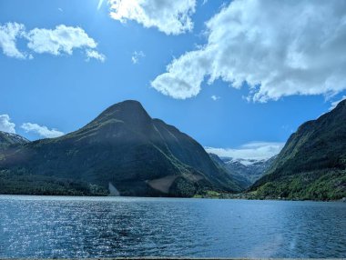 Kayalık kıyı şeridi bulutlu bir gökyüzünün altındaki büyük yeşil dağlarla çevrili sakin bir göle doğru ilerliyor..