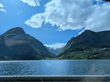 Kayalık kıyı şeridi bulutlu bir gökyüzünün altındaki büyük yeşil dağlarla çevrili sakin bir göle doğru ilerliyor..