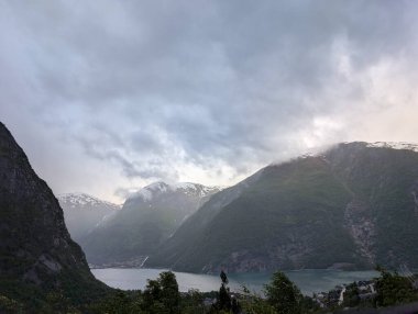 Görkemli fiyort dağları dramatik bir şekilde yükselir, tepelerin arasında göl, bulutlu bir gökyüzünü delip geçen heybetli tepeler