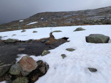 Gri sisli gökyüzünün altındaki kayalık dağ zemininde beyaz kar parçası.