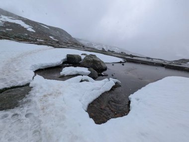 Gri sisli gökyüzünün altındaki kayalık dağ zemininde beyaz kar parçası.