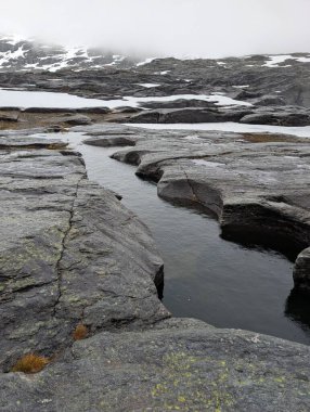 Sarmal su kanalı karanlık, yıpranmış kaya platosuna dönüştü. Şiddetli, bulutlu bir dağ gökyüzünün altında dağınık kar parçacıkları oluştu..