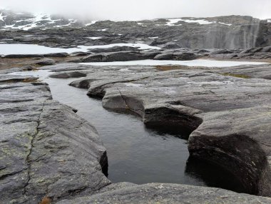 Sarmal su kanalı karanlık, yıpranmış kaya platosuna dönüştü. Şiddetli, bulutlu bir dağ gökyüzünün altında dağınık kar parçacıkları oluştu..