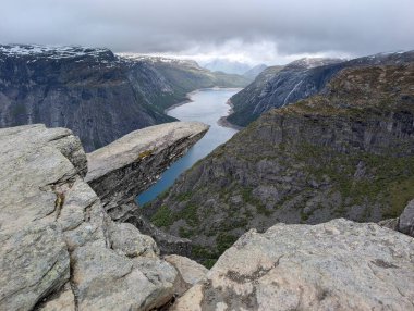 Meşhur Trolltunga kaya oluşumu derin mavi fiyortların üzerinde karamsar bulutlu bir gökyüzünün altında sarp kayalıklarla sarsılıyor..