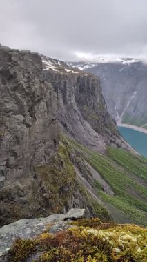 Dikey gri kaya kayalıkları dik yeşil dağ yamacının üzerinden derin vadideki turkuaz göle doğru iniyor.