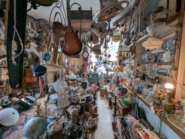 Yoğun bir antika dükkanının iç manzarası, müzik aletleri, klasik lambalar ve çeşitli koleksiyonlarla dolu raflarla çevrili dar koridorları gösteriyor..