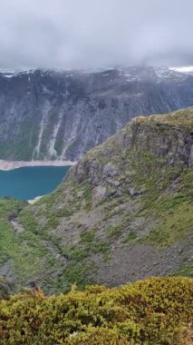 Yüksek irtifa perspektifi ön planda yosun kaplı engebeli bir dağ zirvesini yakalar ve derin, sarp bir vadiye kurulmuş canlı bir turkuaz göle doğru dik bir şekilde düşer..