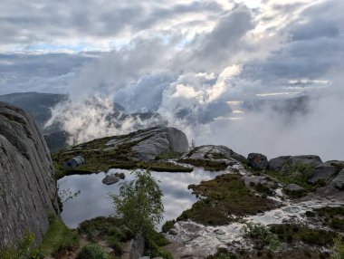 High-angle view reveals a small, calm pool of water reflecting a cloudy sky on a rocky mountain plateau shrouded in mist.