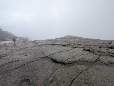 Düşük açılı görüş, geniş, çatlak granit bir platoya bakar. Yoğun beyaz bir sis tabakası içinde, kırmızı ceketli bir kişi uzaklarda yürür..