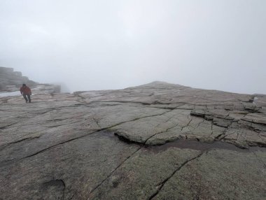 Düşük açılı görüş, geniş, çatlak granit bir platoya bakar. Yoğun beyaz bir sis tabakası içinde, kırmızı ceketli bir kişi uzaklarda yürür..