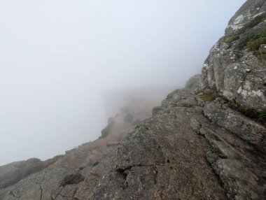 A massive grey granite cliff with a grassy edge and a large boulder is heavily shrouded in thick white mist.