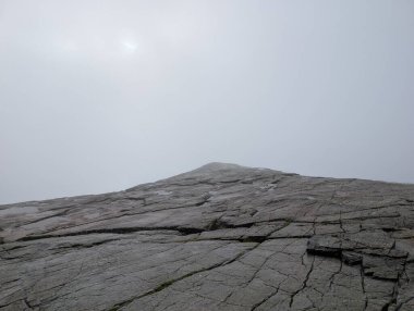 Alçak açılı manzara, yoğun beyaz bir sis tabakasıyla örtülmüş engin, çatlamış granit bir platoya bakar. Güneş sislerin arasından loş bir şekilde görünür..