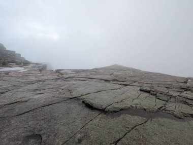 Alçak açılı manzara, yoğun beyaz bir sis tabakasıyla örtülmüş engin, çatlamış granit bir platoya bakar. Güneş sislerin arasından loş bir şekilde görünür..