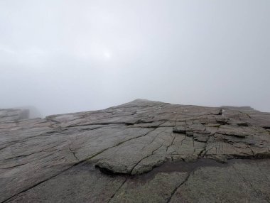Alçak açılı manzara, yoğun beyaz bir sis tabakasıyla örtülmüş engin, çatlamış granit bir platoya bakar. Güneş sislerin arasından loş bir şekilde görünür..