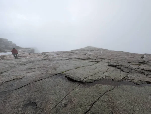 Düşük açılı görüş, geniş, çatlak granit bir platoya bakar. Yoğun beyaz bir sis tabakası içinde, kırmızı ceketli bir kişi uzaklarda yürür..