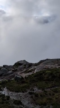 Esrarengiz bir dağ manzarası, bulutlu bir gökyüzünün altında yoğun, girdap gibi beyaz bir sisle kısmen gizlenmiş sivri tepelere doğru uzanan dolambaçlı bir taş yolu içerir..