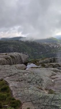 Esrarengiz bir dağ manzarası, bulutlu bir gökyüzünün altında yoğun, girdap gibi beyaz bir sisle kısmen gizlenmiş sivri tepelere doğru uzanan dolambaçlı bir taş yolu içerir..