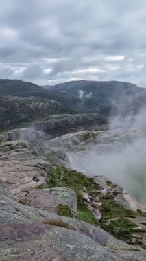 Esrarengiz bir dağ manzarası, bulutlu bir gökyüzünün altında yoğun, girdap gibi beyaz bir sisle kısmen gizlenmiş sivri tepelere doğru uzanan dolambaçlı bir taş yolu içerir..