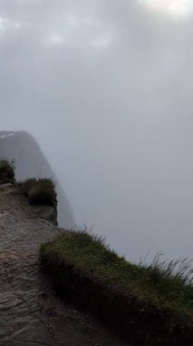 Esrarengiz bir dağ manzarası, bulutlu bir gökyüzünün altında yoğun, girdap gibi beyaz bir sisle kısmen gizlenmiş sivri tepelere doğru uzanan dolambaçlı bir taş yolu içerir..