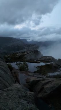 Huysuz, yüksek irtifa manzarası alçak dumanlı, ağır mavi tonlu bir gökyüzünün altında durgun bir dağ havuzu çevreleyen karanlık ve pürüzlü kaya oluşumlarını yakalar..