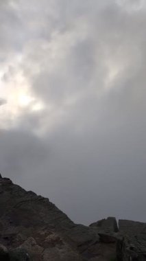 Kasvetli, bulutlu bir gökyüzü kalın, gri bulutlarla doludur. Bu bulutlar güneşi kısmen karartır. Kayalık bir dağın tepesinde yumuşak ve dağınık bir ışık yaratır..