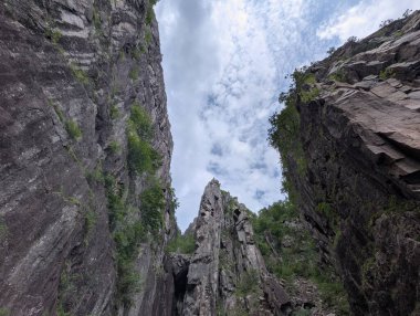 Düşük açı, yüksek gri granit kayalıkları ve yemyeşil bitki örtüsünün parlak, bulutlu bir gökyüzüne doğru yükseldiğini gösteriyor..