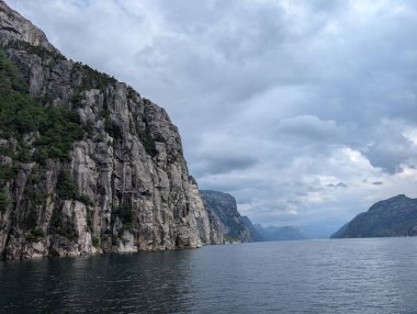Büyük gri granit kayalıklar, seyrek yeşil bitki örtüsü, bulutlu bir gökyüzünün altındaki karanlık sudan dik bir şekilde yükseliyor..