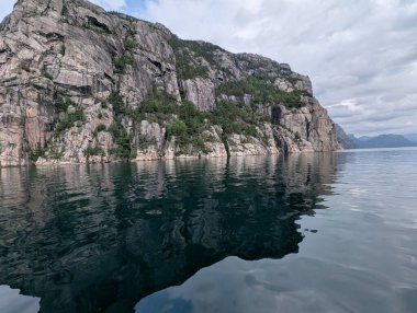 Büyük gri granit kayalıklar, seyrek yeşil bitki örtüsü, bulutlu bir gökyüzünün altındaki karanlık sudan dik bir şekilde yükseliyor..