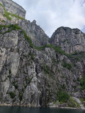 Parlak gri granit kayalıklar, seyrek yeşil bitki örtüsü, bulutlu bir gökyüzünün altındaki karanlık sudan dik bir şekilde yükseliyor.