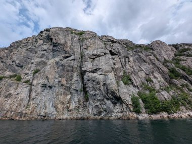 Karanlık suların üzerinde bulutlu bir gökyüzü beliriyor, bu sarp, gri granit kayalıklardan yeşil bitki örtüsü seyrek olarak yükseliyor..